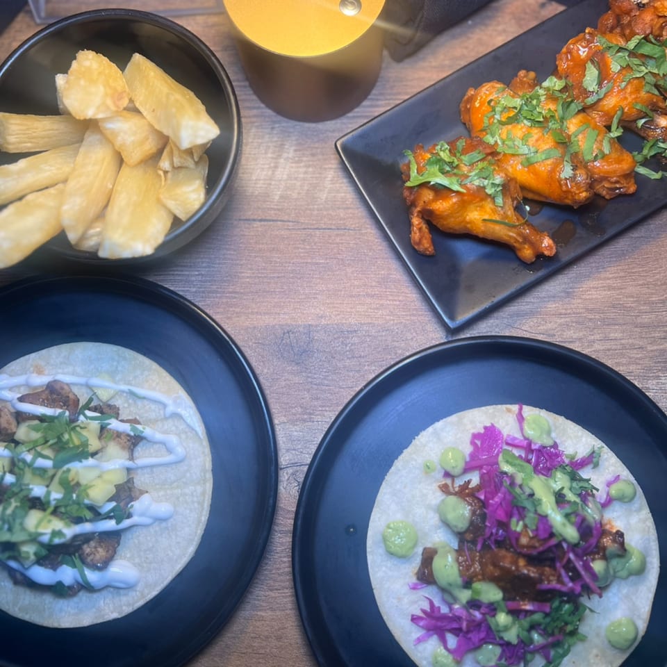 Two tacos, a bowl of yuca fries, and chicken wings on a wooden table at The Taco Bar.