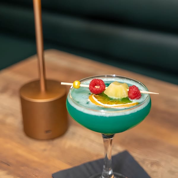 Vibrant blue cocktail garnished with raspberries, pineapple, and orange slice on a wooden table.