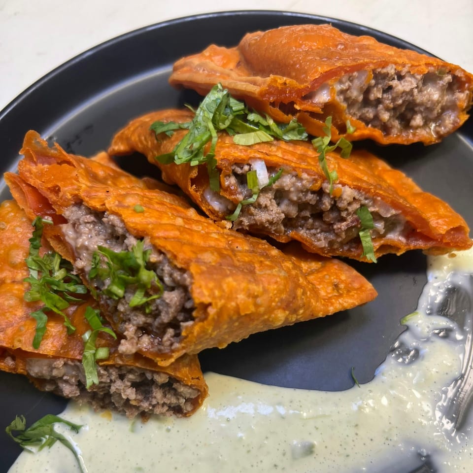 Close-up of several crispy, orange-hued empanadas filled with ground beef and topped with cilantro, served with a creamy green sauce.