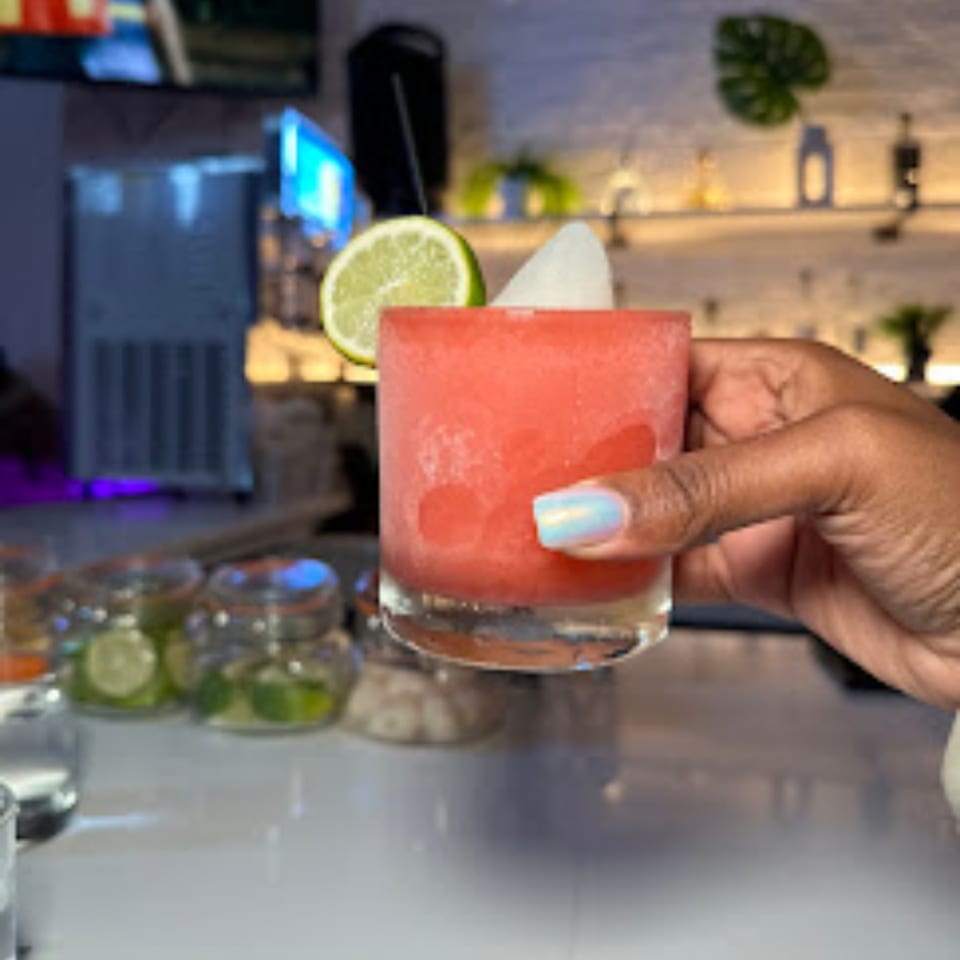 A hand holding a frosted glass of red margarita garnished with a lime slice and white fruit.