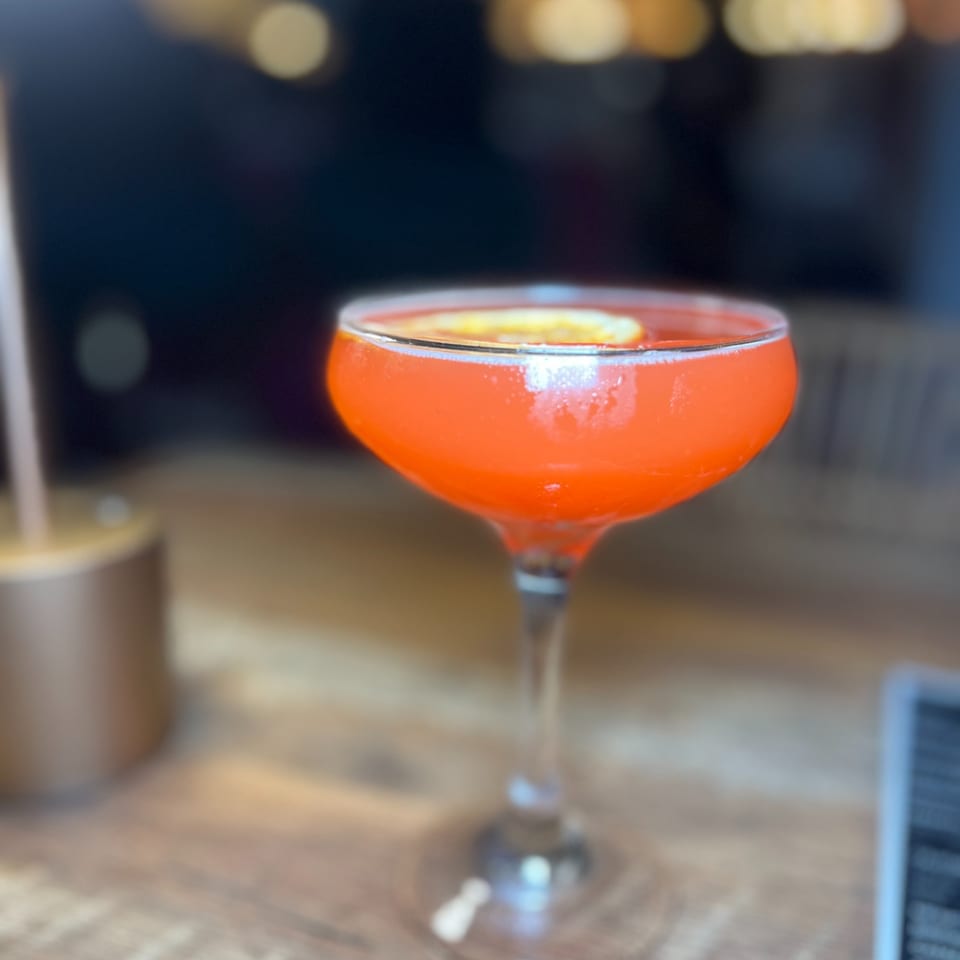A vibrant red cocktail with a citrus slice, served in a coupe glass on a wooden table.
