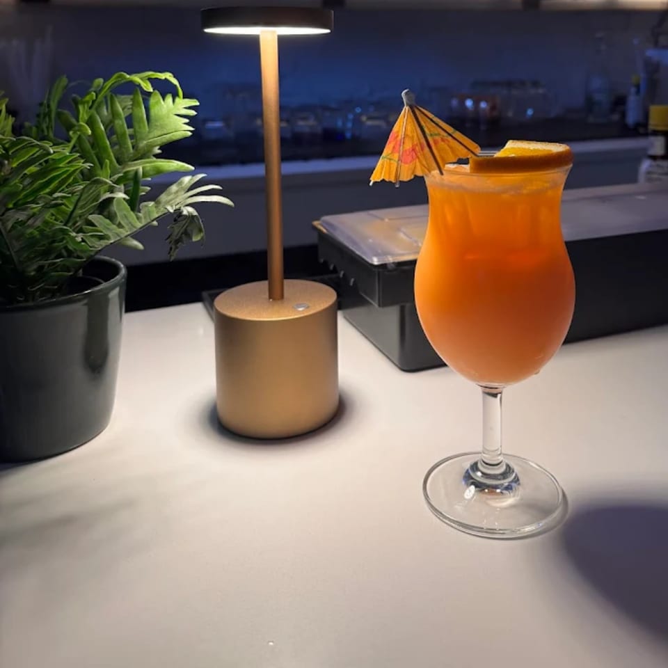 A vibrant orange cocktail with a paper umbrella and orange slice, next to a plant and lamp.