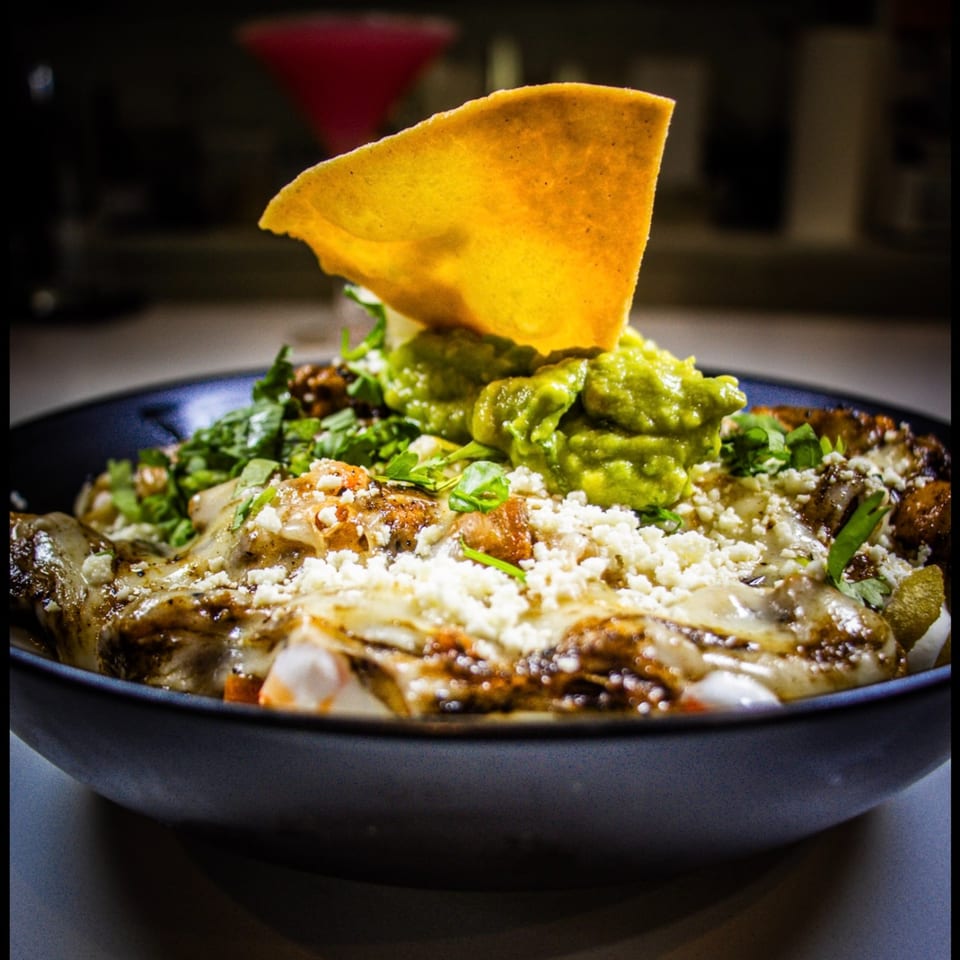 A bowl of The Taco Bar's Mexican food, topped with guacamole, cheese, and a tortilla chip.